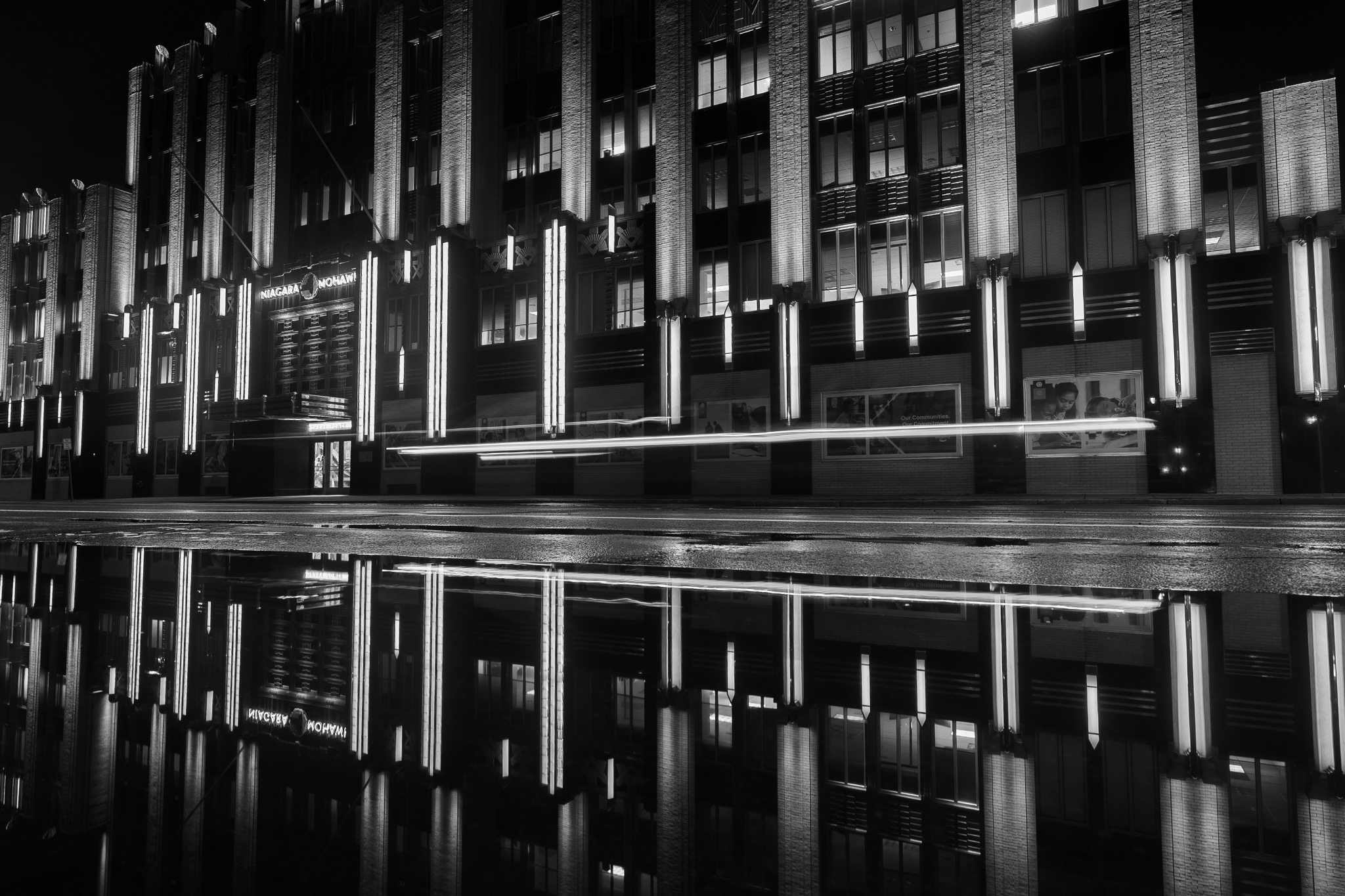 Illuminated Niagara Mohawk facade, a passing light trail and a puddle reflection in black and white