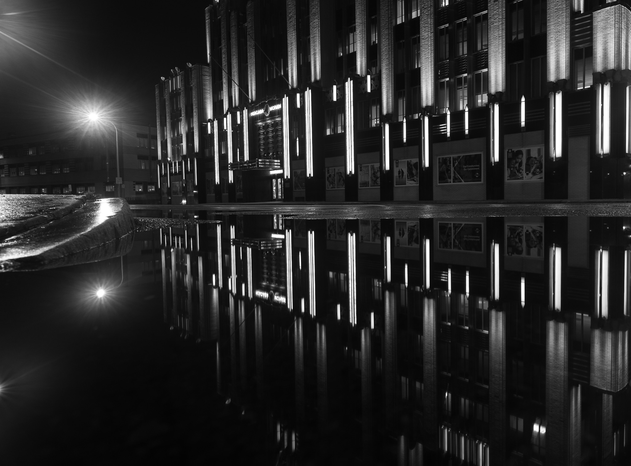 Low black-and-white view of an illuminated facade and streetlamp reflected beside a curved curb