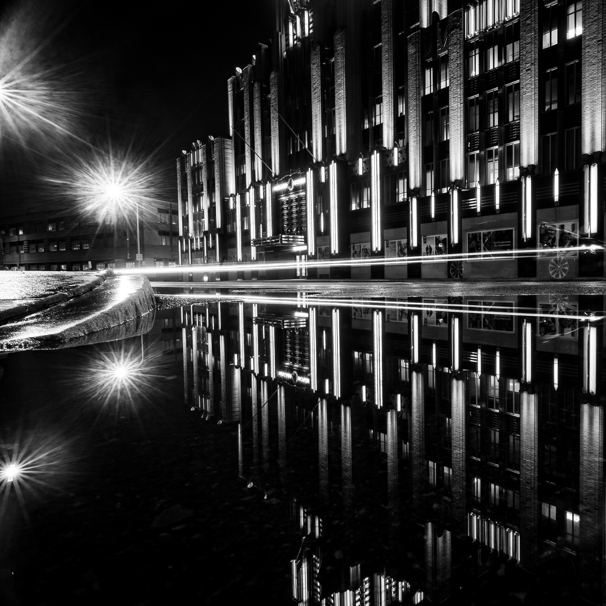 Square black-and-white night scene with starburst lamps, traffic light trails and reflected architecture