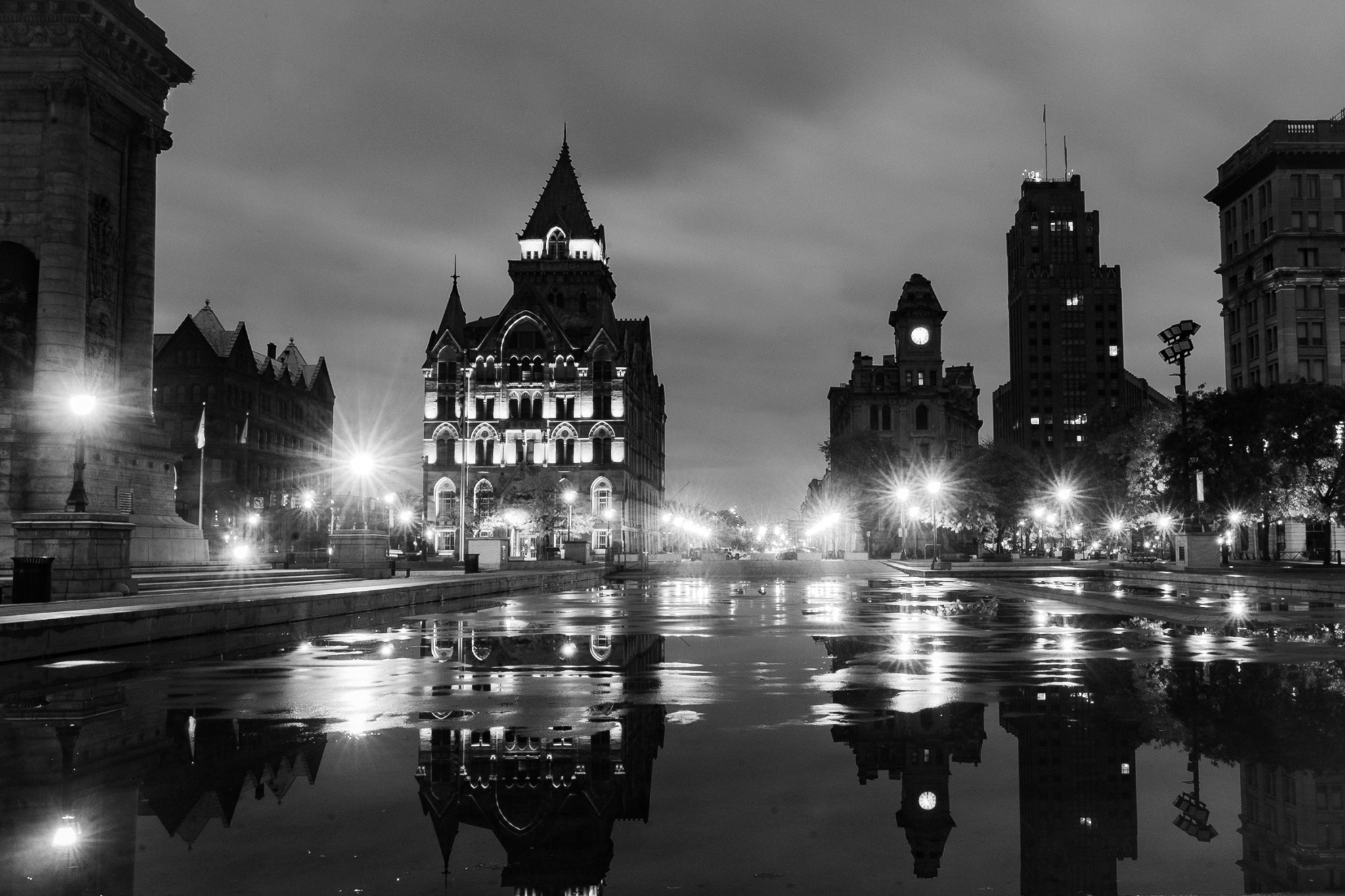 Historic city buildings, a clock tower and bright streetlamps reflected across wet pavement in black and white